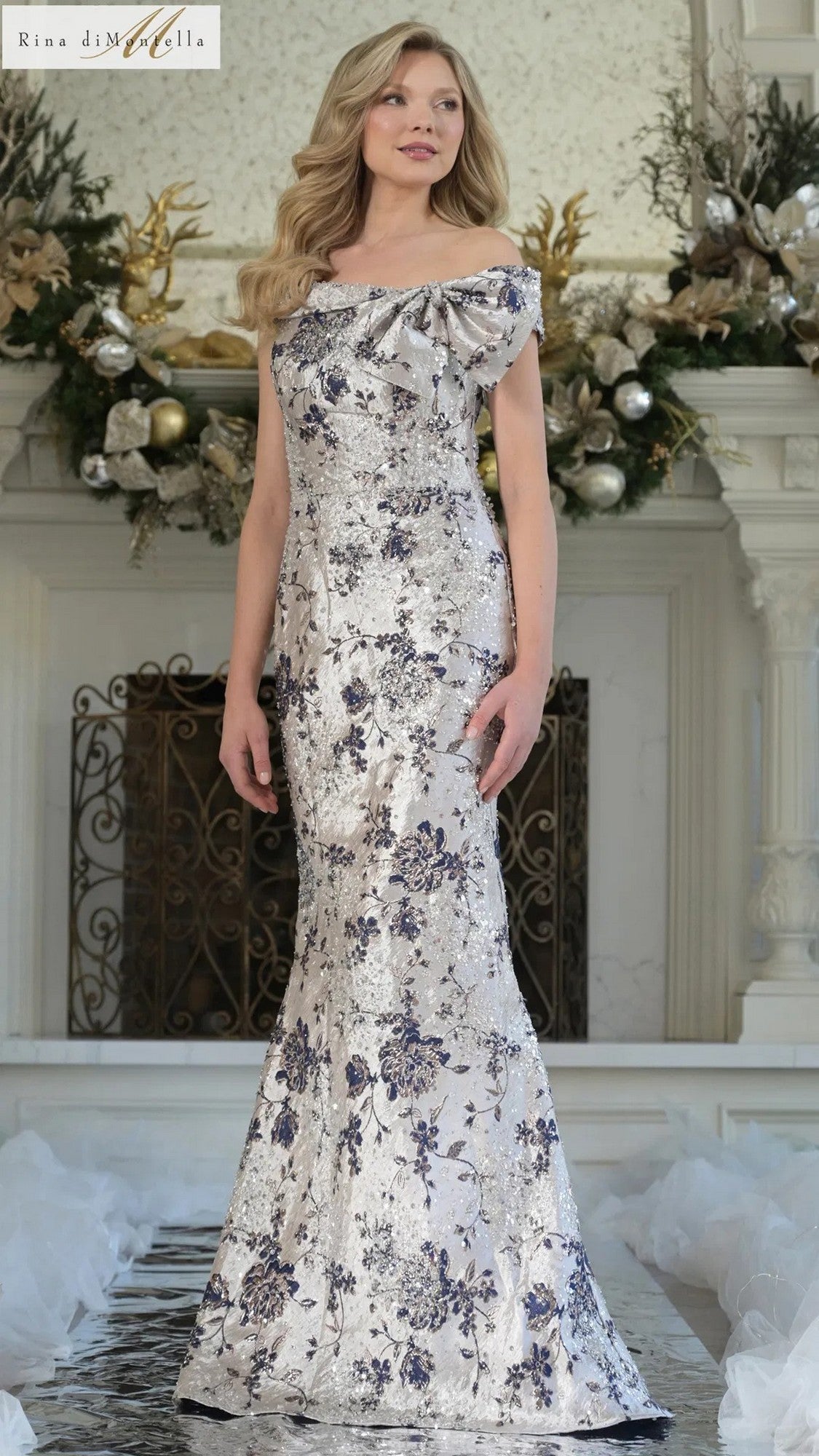 A woman stands indoors wearing the Rina Di Montella RD3106 long formal dress in Navy Multi, featuring blue floral patterns and a subtle mermaid silhouette. Festive decorations and white fabric pieces appear in the background.